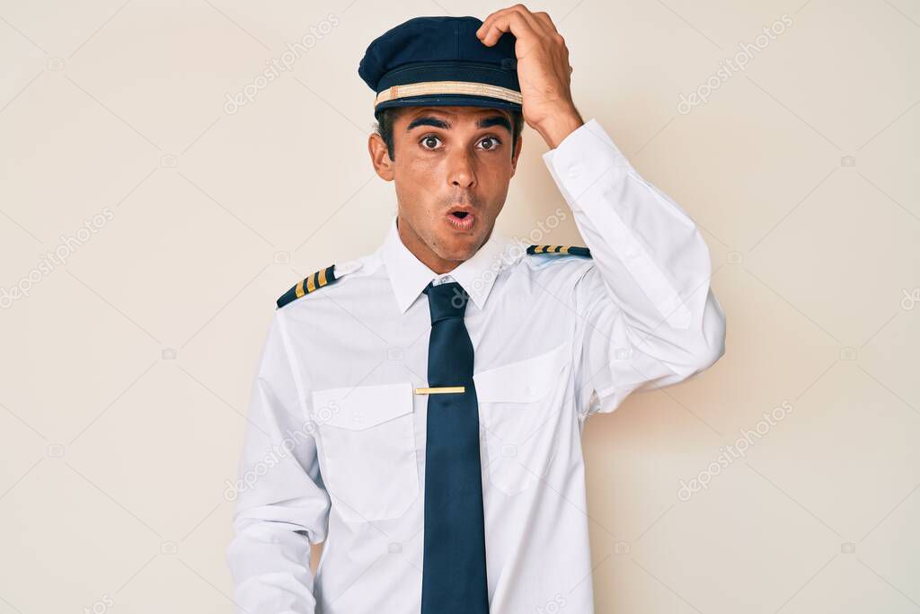 Young hispanic man wearing airplane pilot uniform surprised with hand on head for mistake, remember error. forgot, bad memory concept. 