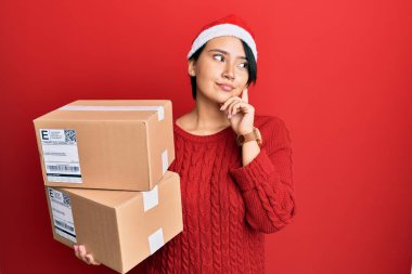 Kısa saçlı, Noel şapkası takan güzel bir kadın teslimat paketleri taşıyan ciddi bir yüz ifadesi olan eli çenesinde soru soran, kafa karıştırıcı bir fikir hakkında düşünceli bir kadın. 