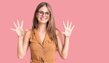 Genç, güzel, sarışın, günlük kıyafetler ve gözlük takan, on numaralı parmaklarıyla işaret eden, kendinden emin ve mutlu bir şekilde gülümseyen bir kadın.. 