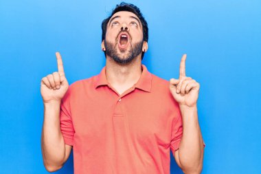 Gündelik giysiler giyen genç İspanyol adam şaşırmış ve şaşırmış bir şekilde yukarı bakıyor ve parmaklarıyla işaret ediyor ve kollarını kaldırıyor.. 