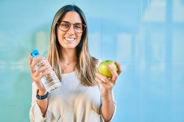 Genç beyaz kadın gülümsüyor. Şehirde yeşil elma ve bir şişe su tutuyor..