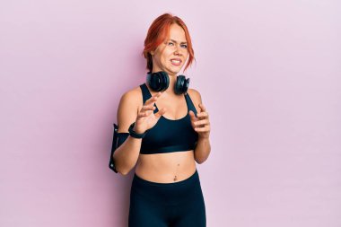 Genç, kızıl saçlı, spor kıyafetleri giyen, kulaklık takan, tiksindirici bir yüz ifadesi takan, tiksindirici bir yüz ifadesi takınan genç bir kadın. Eller havada. 