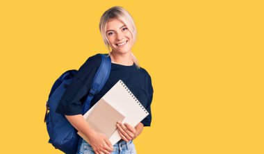 Genç, sarışın, öğrenci çantası taşıyan, elinde not defteri olan güzel bir kadın. Pozitif ve mutlu görünüyor. Dişlerini gösteren bir gülümsemeyle ayakta duruyor. 