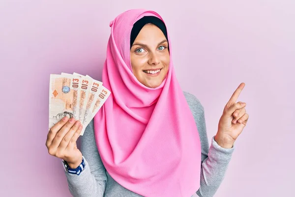 年轻的高加索女人头戴伊斯兰式头巾 手里拿着10磅钞票 高兴地微笑着 手指手画脚地指向旁边 — 图库照片
