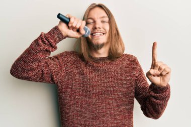 Uzun saçlı yakışıklı beyaz adam mikrofon kullanarak şarkı söylüyor. Bir fikir veya soru işareti ile gülümsüyor. 