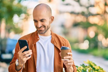 Genç Latin kökenli kel adam akıllı telefon kullanıyor. Şehirde kahve içiyor..