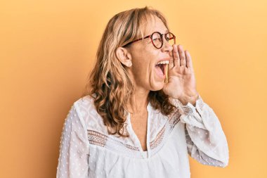 Sarışın, orta yaşlı bir kadın sarı arka plan üzerine gündelik kıyafetler giyip el ele verip bağırıyor. iletişim konsepti. 