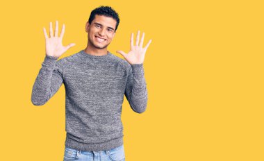 İspanyol asıllı yakışıklı genç adam gündelik süveter giyer ve parmaklarıyla on numarayı gösterirken kendinden emin ve mutlu bir şekilde gülümser.. 