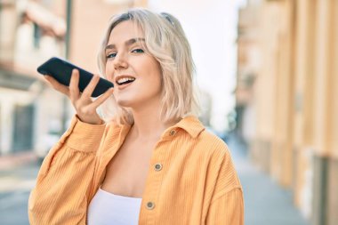 Genç sarışın kız gülümseyerek şehirde akıllı telefon kullanarak sesli mesaj gönderiyor..