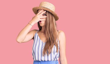 Genç, sarışın, yaz şapkası takan, şokla yüzünü ve gözlerini kapatan, utangaç ifadesiyle parmaklarının arasından bakan genç bir kadın.. 