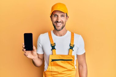 Tamirci üniforması giyen yakışıklı genç adam akıllı telefon ekranı gösteriyor. Pozitif ve mutlu görünüyor. Kendine güvenen bir gülümsemeyle dişlerini gösteriyor. 