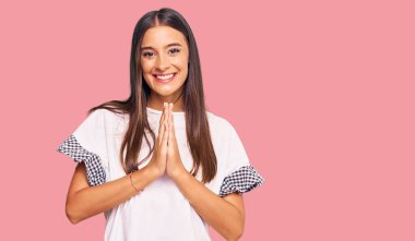 Gündelik giysiler giyen genç İspanyol kadın elleriyle dua ediyor af diliyor kendinden emin bir şekilde gülümseyerek.. 