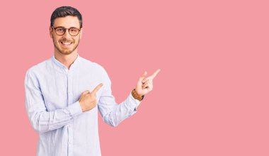 Zarif iş gömleği ve gözlüklü yakışıklı genç adam gülümsüyor ve kameraya bakıyor iki eliyle ve parmaklarıyla kenarı işaret ediyor.. 