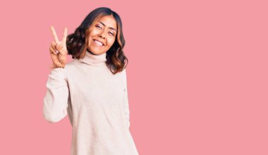 Balıkçı yaka kazak giyen, gülümseyen, parmaklarını zafer işareti yaparken gösteren genç ve güzel bir ırk kadını. İki numara.. 