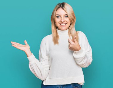 Kış süveteri giyen genç beyaz kadın avuç içi gösteriyor ve baş parmaklarıyla iyi bir hareket yapıyor, mutlu ve neşeli gülümsüyor. 