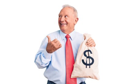 Yakışıklı, beyaz saçlı, şık kıyafetler giyen, para çantası tutan, başparmağıyla yan tarafı gösteren, ağzı açık gülümseyen bir adam. 