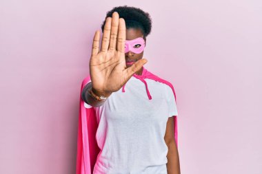 Genç Afrikalı Amerikalı kız süper kahraman maskesi ve pelerin giyip avuç içiyle şarkı söylemeyi kesiyor. Yüzünde olumsuz ve ciddi bir jest olan uyarı ifadesi. 