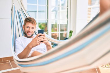 Yakışıklı beyaz adam mutlu bir şekilde gülümsüyor terasta dinlenirken akıllı telefon kullanıyor.