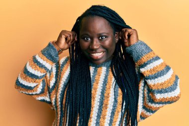 Saçları örgülü, sıradan bir süveter giyen, kulakları parmaklarıyla çeken genç siyah bir kadın. Komik bir jest. deneme sorunu 
