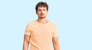 Kıvırcık saçlı, sıradan kıyafetler giyen, yüzünde ciddi bir ifade olan genç yakışıklı bir adam. Basit ve doğal bir şekilde kameraya bakıyorsun.. 