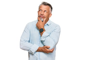 Orta yaşlı, gri saçlı, günlük kıyafetler giyen, bir soru hakkında endişelenen, eli çenesinde endişeli ve gergin bir adam. 