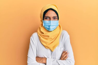 Geleneksel İslami tesettür ve tıbbi maske takan, kollarını kavuşturup kameraya gülümseyen genç esmer Arap kadın. pozitif kişi. 