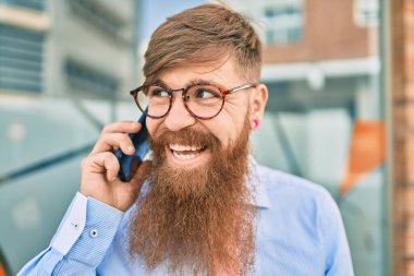 Genç kızıl saçlı işadamı, mutlu bir şekilde gülümsüyor ve şehirde duran akıllı telefonda konuşuyor..
