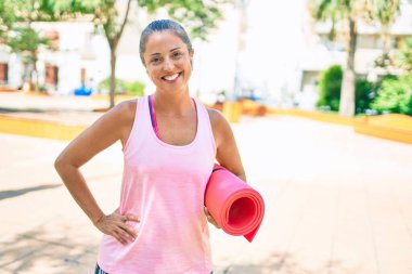 Orta yaşlı sporcu kadın parkta yoga minderi tutarken mutlu gülümsüyor.