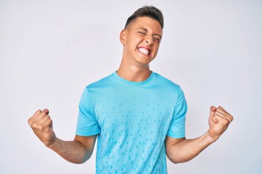 Genç İspanyol çocuk gündelik kıyafetler giyip çok mutlu ve heyecanlı bir şekilde kollarını kaldırıp, gülümseyerek ve başarı için bağırarak kazanan jestler yapıyor. kutlama konsepti. 