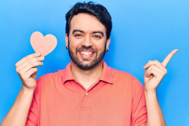 Elinde kalp şeklinde kâğıt tutan genç İspanyol adam gülümsüyor mutlu bir şekilde elini ve parmağını yana doğru gösteriyor. 
