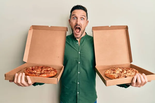 Handsome man with beard holding two italian pizzas celebrating crazy and amazed for success with open eyes screaming excited.