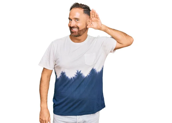 噂やゴシップに耳を傾ける耳の上に手で笑みを浮かべてカジュアルなネクタイ染料のTシャツを着てハンサムな中年の男性 聴覚障害の概念 — ストック写真