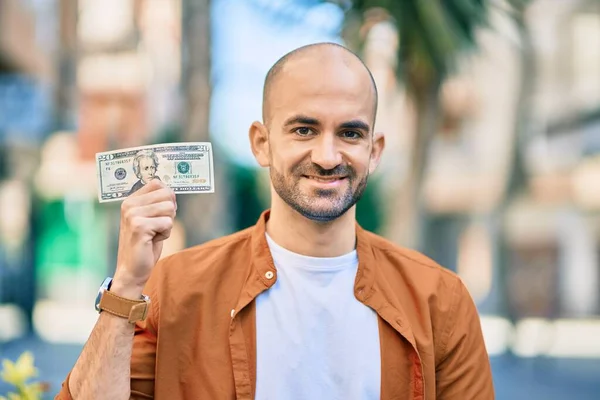 Young hispanic bald man smiling happy holding one usa dollar at the city.