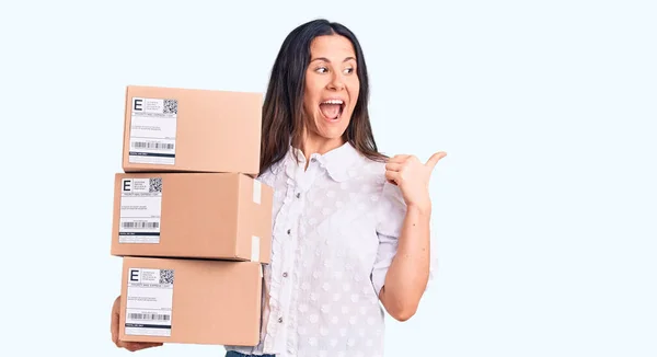 Young Beautiful Brunette Woman Holding Delivery Cardboard Package Pointing Thumb — Stock Photo, Image