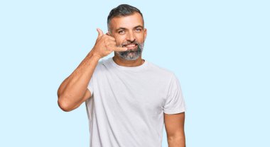 Orta yaşlı, beyaz tişört giyen, gülümseyen, el ve parmaklarıyla telefon hareketi yapan, telefonda konuşan yakışıklı bir adam. iletişim kavramları. 