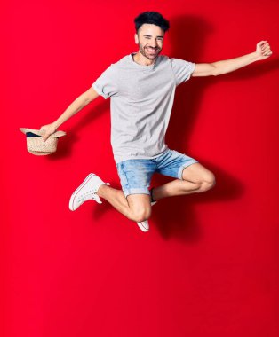 Genç, yakışıklı, sıradan kıyafetler giyen, mutlu gülümseyen bir İspanyol. Yüzünde tebessümle zıplayarak yaz şapkası tutarak izole edilmiş kırmızı arka planda yumruğunu havaya kaldırarak kutluyor..