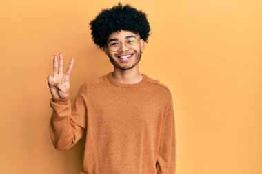Afro saçlı, sıradan bir kazak giyen genç Afrikalı Amerikalı üç numarayı gösterip gülümserken kendinden emin ve mutlu bir şekilde işaret ediyor.. 