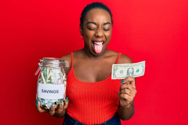 Genç Afro-Amerikalı kadın elinde kavanozla 1 dolarlık banknot tutarken mutlu bir ifadeyle dilini dışarı çıkarıyor.. 
