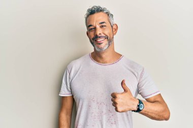 Orta yaşlı, gri saçlı, günlük kıyafetler giyen, mutlu mesut el hareketi yapan bir adam. Kameraya bakıp başarıyı gösteren ifade. 