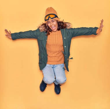 Kışlık giysiler giyen genç, güzel Latin kadın mutlu gülümsüyor. Yüzünde ve kollarında tebessümle, izole edilmiş sarı arka planda atlıyor.