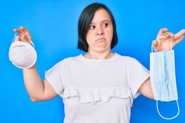 Down sendromlu esmer kadın elinde iki farklı güvenlik maskesi ve kafası karışık bir ifade tutuyor. şüphe kavramı. 