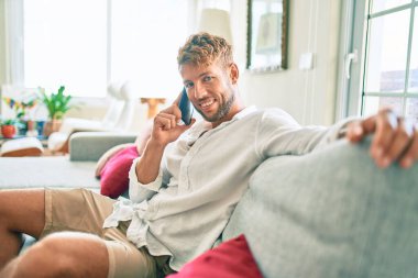 Yakışıklı beyaz adam gülümsüyor. Evdeki kanepede oturup telefonla konuşuyor.