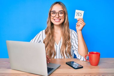 Genç ve güzel sarışın bir kadın elinde evet hatırlatıcısıyla masada oturmuş pozitif ve mutlu görünüyor ve kendine güvenen bir gülümsemeyle dişlerini gösteriyor. 