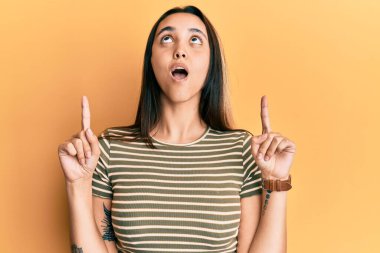 Gündelik çizgili tişört giyen genç İspanyol kadın şaşkın şaşkın yukarı bakıyor ve parmaklarıyla işaret ediyor ve kollarını kaldırıyor.. 
