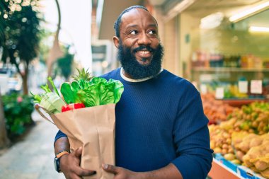 Sakallı Afrikalı Amerikalı bir adam elinde süpermarketten alınmış kağıt poşetler tutuyor.