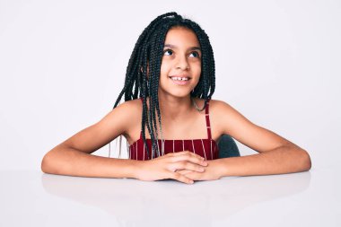 Saçları örgülü genç Afro-Amerikalı kız masaya oturmuş yüzünde gülümseme ve doğal bir ifadeyle diğer tarafa bakıyor. Kendinden emin gülen. 