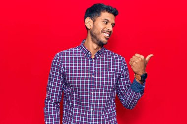 Gündelik giysiler giymiş genç Latin bir adam başparmağını yana kaldırmış, ağzı açık gülümsüyor. 