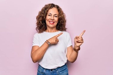 Orta yaşlı güzel bir kadın, günlük tişört giyip pembe arka planda dikiliyor gülümsüyor ve kameraya bakıyor iki eliyle ve parmaklarıyla yan tarafa bakıyor..