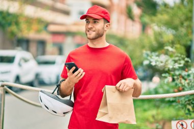 Beyaz atlı genç teslimatçı akıllı telefon kullanıyor ve şehirde kağıt torba taşıyor..