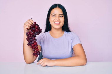 Genç, güzel Asyalı kız elinde taze üzümlerden bir dal tutuyor. Pozitif ve mutlu görünüyor. Kendine güvenen bir gülümsemeyle dişlerini gösteriyor. 